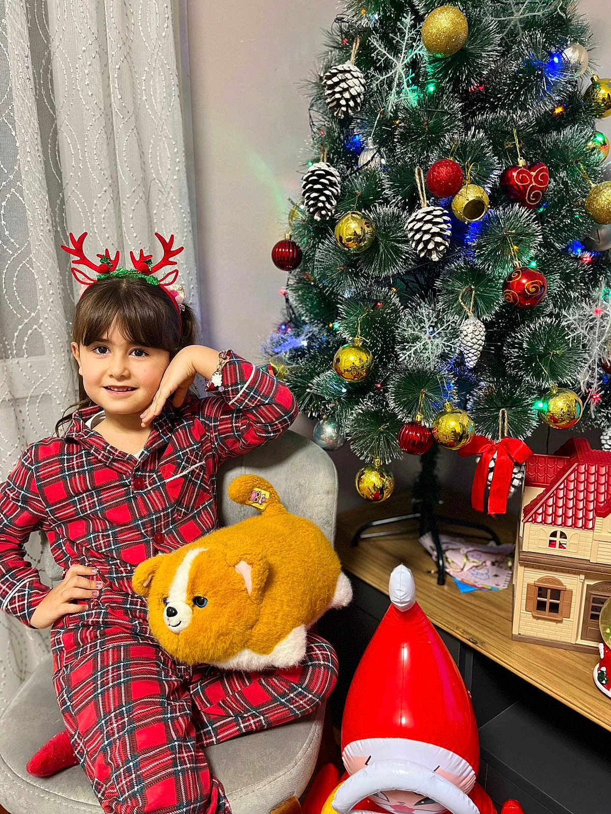 Shams is registered to the contest to win money with this photo: child, girl, smile, pajamas, plaid, reindeer_headband, plush_toy, stuffed_animal, christmas_tree, ornament, pinecone, bauble, christmas_decor, chair, curtain, indoor, holiday, festive, toy_house, santa_figure
