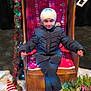 child, winter_clothing, hat, jacket, chair, wooden_throne, christmas_lights, garland, artificial_snow, holiday_decoration, indoor, smiling, rollerblades, floor, festive, red_pattern, white_hat, pink_shoes, greenery, cardboard_santa