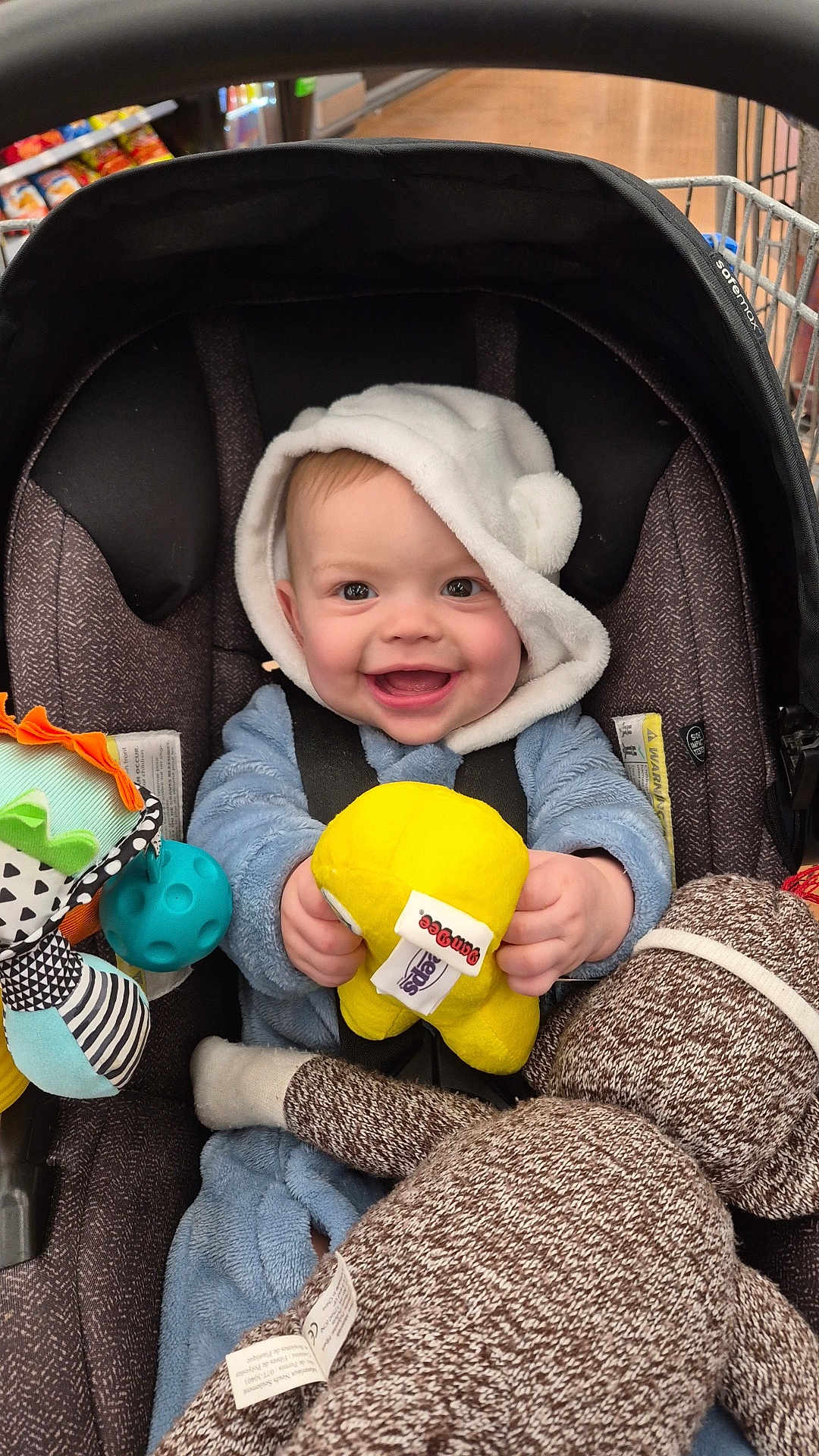 Zayvien is registered to the contest to win money with this photo: baby, infant, stroller, car_seat, toy, plush_toy, stuffed_animal, smile, happy, hood, blue_outfit, seatbelt, shopping_cart, supermarket, portrait, closeup, cheerful, yellow_toy, sock_monkey, hand