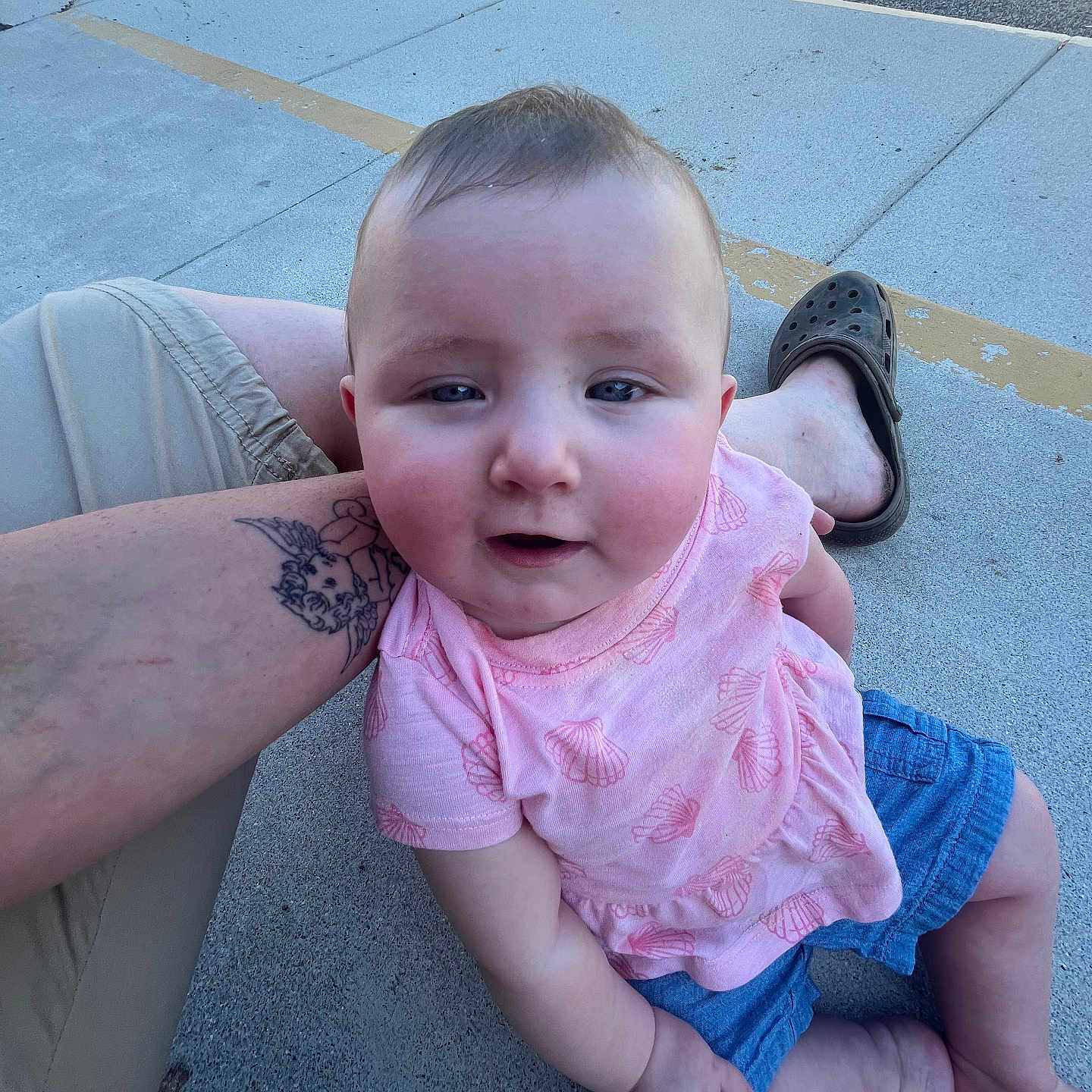 Peyton is registered to the contest to win money with this photo: baby, child, infant, sitting, portrait, outdoor, sunlight, pink_shirt, blue_shorts, smiling, rosy_cheeks, short_hair, face, person, adult_arm, tattoo, crocs, concrete, playground, casual_photo