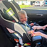 baby, child, car, car_seat, pacifier, sitting, smiling, blond_hair, outdoor, sunlight, vehicle, passenger, seatbelt, infant, toy, stuffed_animal, suburban_house, window, road_trip, portrait