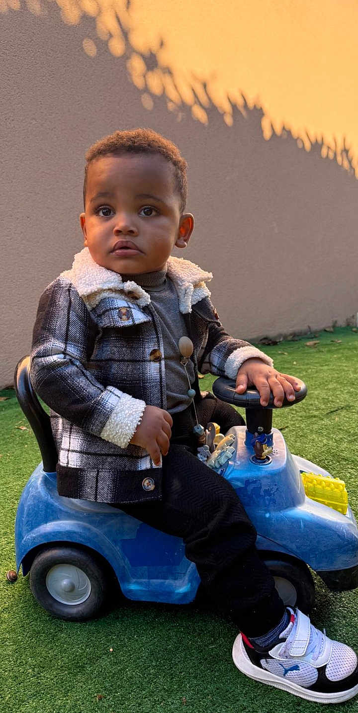 Ozani participe au concours pour gagner de l'argent avec cette photo : toddler, child, toy_car, outdoor, grass, jacket, plaid, shoes, sneakers, curious, young_child, person, casual_clothing, playtime, daylight, shadow, wall, greenery, seat, hands