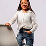 child, girl, glasses, white_blouse, jeans, ripped_jeans, chair, long_hair, casual_clothing, standing, smile, portrait, studio, fashion, modern_chair, hand_in_pocket, barefoot, confident, young_person, indoor