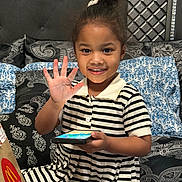 Ashley is registered to the contest to win money with this photo: child, smiling, wave, smartphone, striped_dress, bed, pillow, headband, hair_bun, indoor, happy, mcdonalds_bag, patterned_bedding, portrait, young_girl, hand, sitting, black_and_white_stripes, decorative_headboard, cozy