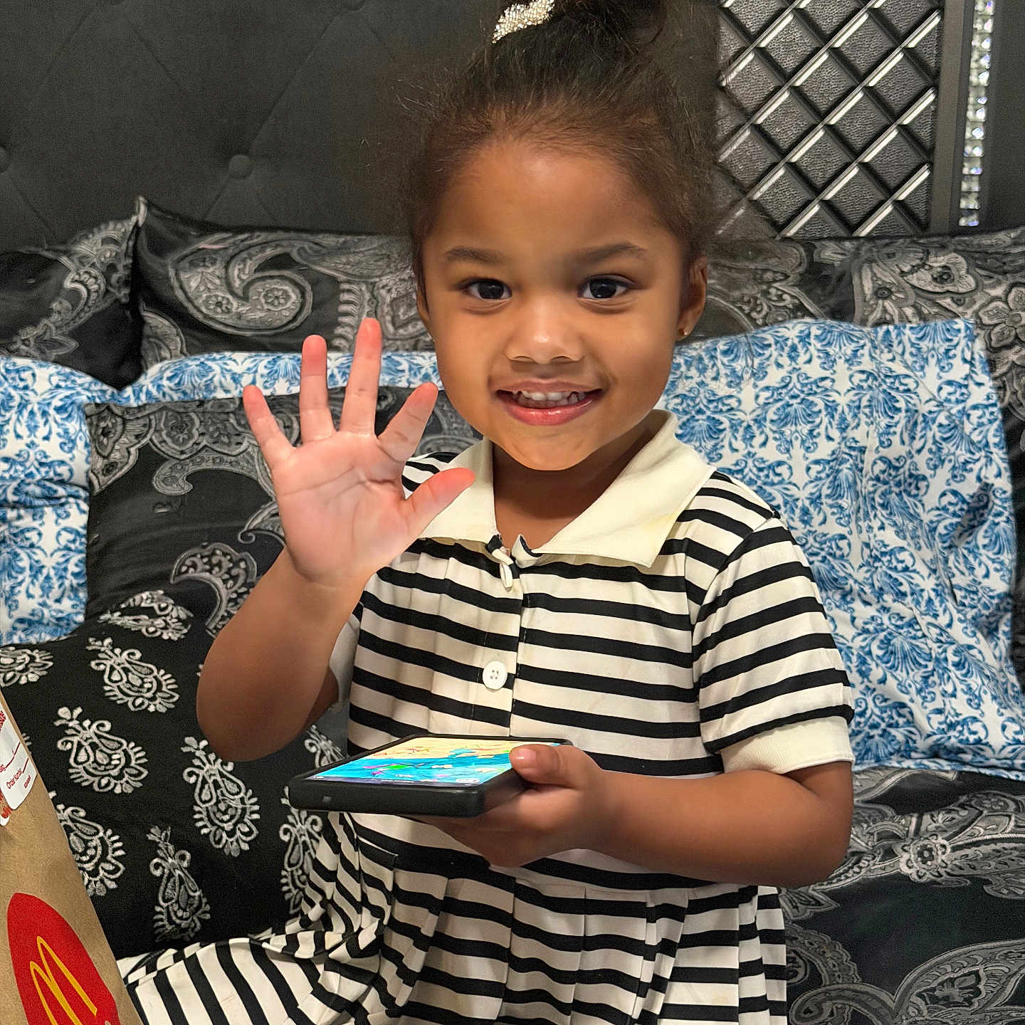 Ashley is registered to the contest to win money with this photo: bed, black_and_white_stripes, child, cozy, decorative_headboard, hair_bun, hand, happy, headband, indoor, mcdonalds_bag, patterned_bedding, pillow, portrait, sitting, smartphone, smiling, striped_dress, wave, young_girl