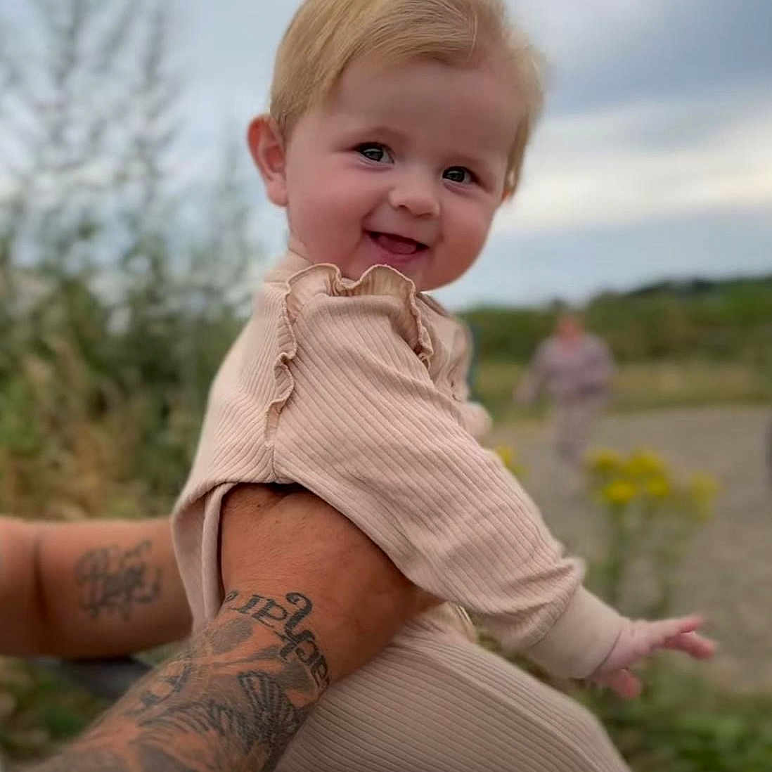 Ava-Gray is registered to the contest to win money with this photo: arm, baby, blonde_hair, blurred_background, child, clothing, cloudy_sky, cute, greenery, happy, holding, infant, nature, outdoor, person, playful, portrait, smiling, tattoo, warm