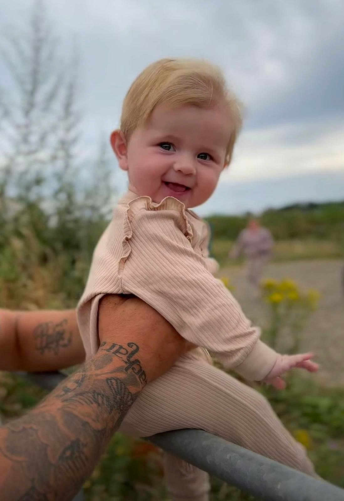 Ava-Gray is registered to the contest to win money with this photo: baby, child, smiling, tattoo, arm, outdoor, nature, cloudy_sky, greenery, person, holding, cute, portrait, happy, infant, clothing, blonde_hair, blurred_background, playful, warm