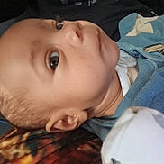 Carson is registered to the contest to win money with this photo: baby, infant, face, closeup, blue_clothing, lying_down, curious, young_child, skin, head, ear, eyes, fabric, blanket, indoors, portrait, person, soft_lighting, child, sleepwear