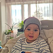 Gabriel a rejoint le concours — aidez-le/la à gagner de superbes lots ! baby, child, striped_clothing, hat, high_chair, blanket, indoor, plant, curtain, window, face, hands, cozy, cute, person, infant, smiling, home, natural_light, seated