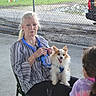 blue_lanyard, casual, chain_link_fence, chair, concrete_floor, conversation, daylight, dog, elderly_woman, event, fluffy_dog, grey_hair, happy_dog, leash, outdoor, park, people, sitting, small_dog, tie_dye_shirt
