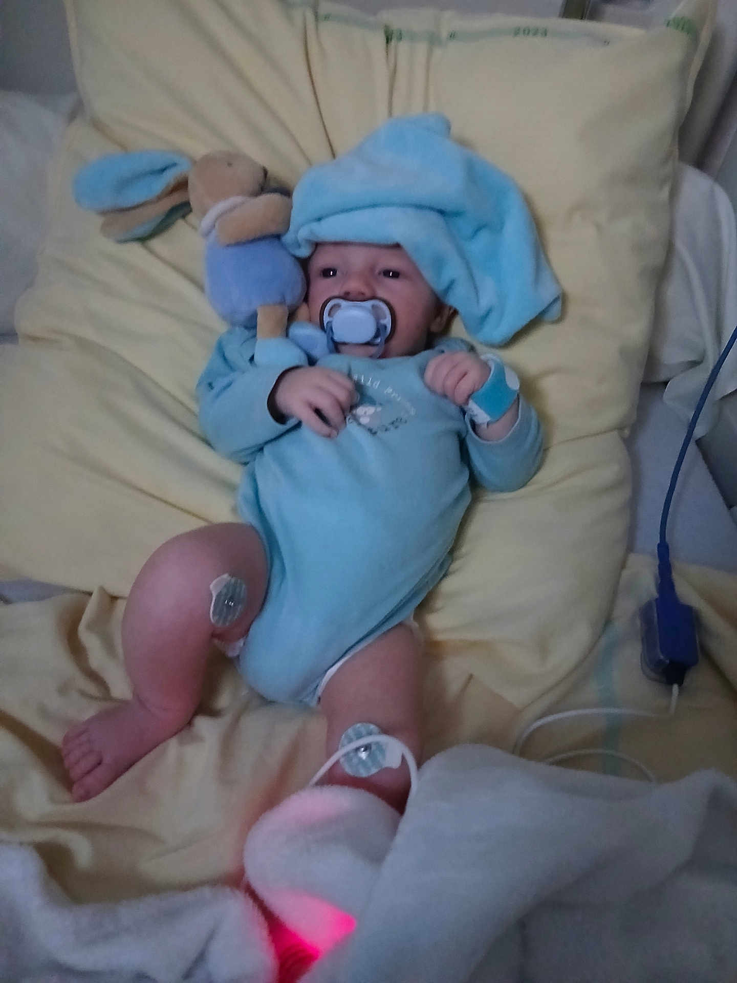 Mayron a rejoint le concours — aidez-le/la à gagner de superbes lots ! baby, pacifier, plush_toy, blue_onesie, blue_hat, pillow, medical_sensor, hospital, infant, bed, blanket, monitoring_device, soft_toys, lying_down, cute, child, person, healthcare, resting, indoors