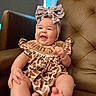 baby, child, smiling, bow, headband, romper, patterned_clothing, leather_chair, happy, portrait, sitting, infant, cute, indoors, furniture, skin, feet, hand, face, person