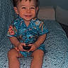 child, boy, toddler, smiling, happy, portrait, sitting, bed, bedroom, indoors, floral_shirt, blue_clothing, shorts, barefoot, remote_control, toy, patterned_blanket, furniture, single_face, photograph