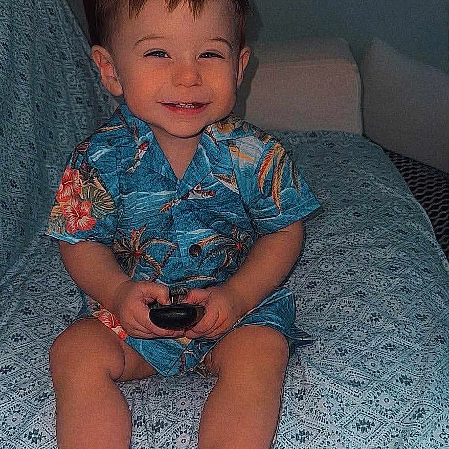 Kaysen is registered to the contest to win money with this photo: child, boy, toddler, smiling, happy, portrait, sitting, bed, bedroom, indoors, floral_shirt, blue_clothing, shorts, barefoot, remote_control, toy, patterned_blanket, furniture, single_face, photograph