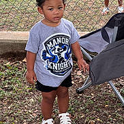 Karia joined the competition — help win amazing prizes! toddler, child, outdoor, grass, leaves, chain_link_fence, tshirt, shorts, sandals, chair, person, curly_hair, top_knot, sidewalk, cheerleading, logo, standing, daylight, expression, casual_clothing