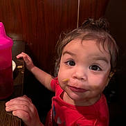 Amari is registered to the contest to win money with this photo: toddler, child, face, hands, food, sippy_cup, table, wood_paneling, red_shirt, curly_hair, indoor, closeup, expression, messy, cute, portrait, person, young_child, sitting, eye_contact