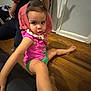 toddler, child, pink_clothing, green_shorts, wooden_floor, indoor, person, arm, shadow, playful, headwear, curious, sitting, casual, home, flooring, young_child, human, portrait, funny_pose