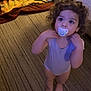 toddler, child, pacifier, curly_hair, barefoot, onesie, carpet, indoor, bed, rug, floor, person, looking_up, standing, home, curious, young_child, baby, furniture, soft_lighting