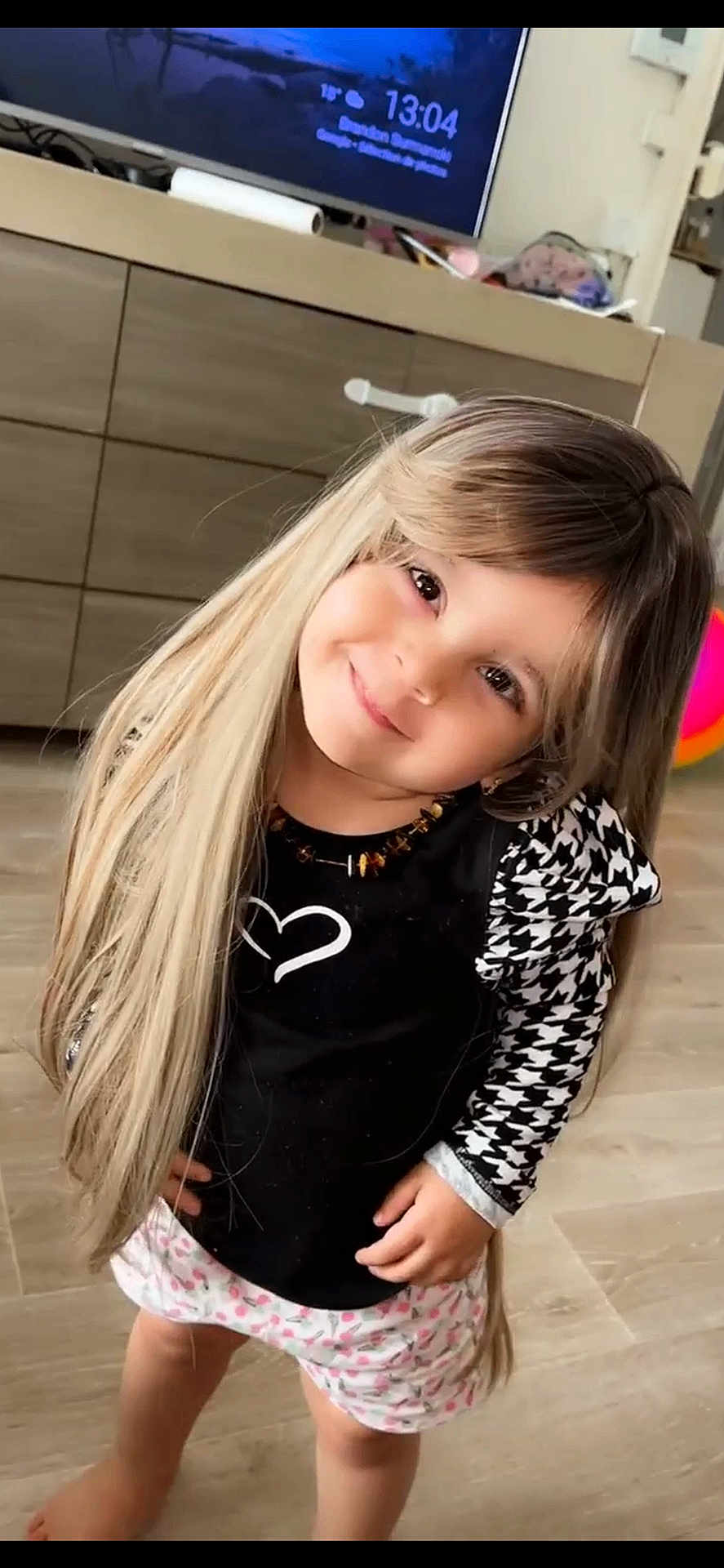 Mélina participe au concours pour gagner de l'argent avec cette photo : child, smiling, wig, long_hair, black_top, patterned_sleeves, shorts, barefoot, wooden_floor, indoor, living_room, furniture, cabinet, television, necklace, cute, portrait, playful, happy, young