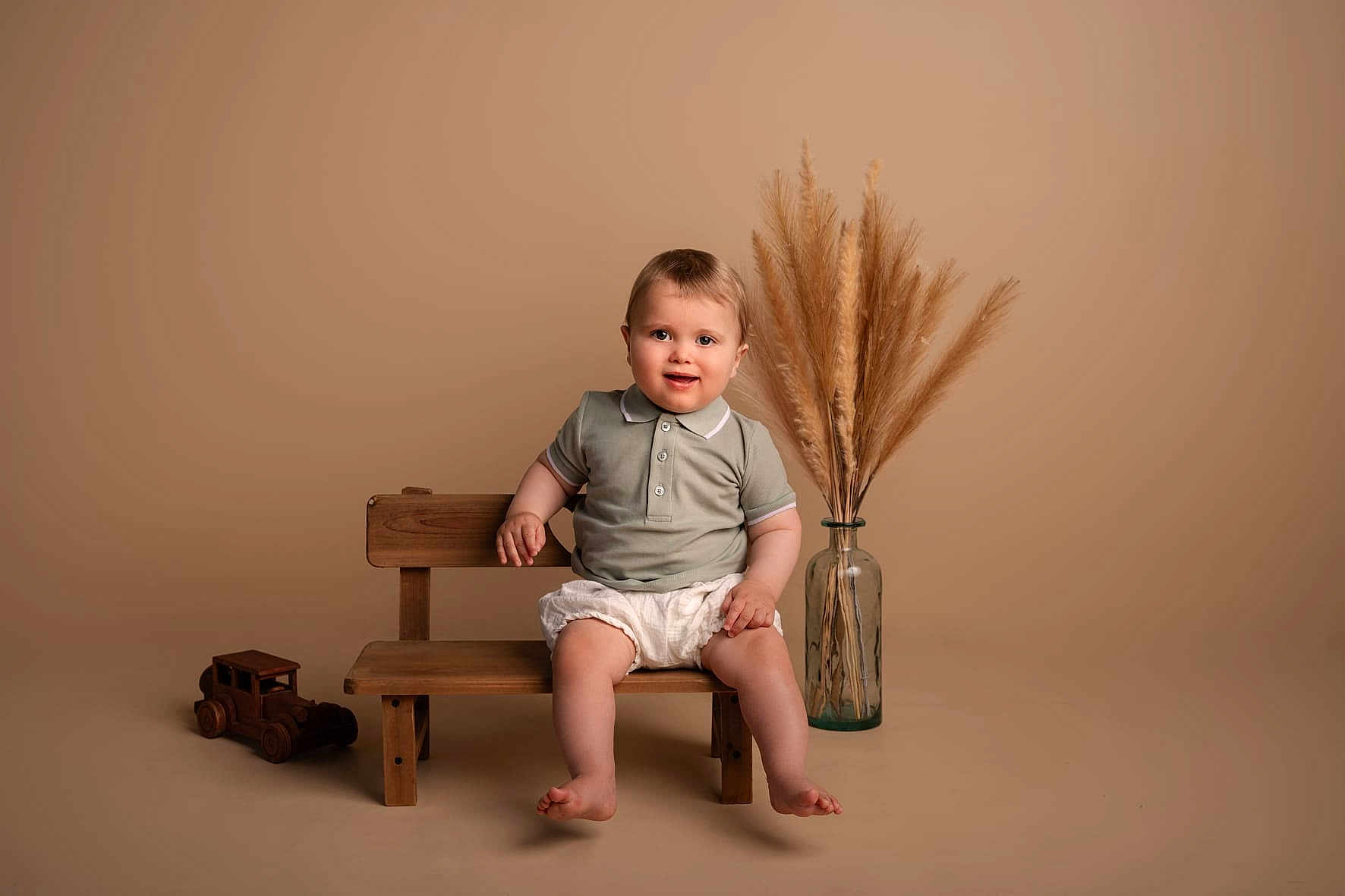 Armand a rejoint le concours — aidez-le/la à gagner de superbes lots ! toddler, child, bench, wooden_bench, toy_car, wooden_toy, pampas_grass, vase, glass_vase, smiling, shorts, polo_shirt, barefoot, indoor, studio, neutral_background, cute, portrait, seated, happy