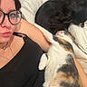 woman, glasses, calico_cat, black_dog, blanket, couch, indoor, pet, relaxing, laying_down, casual, portrait, animal, sleeping_cat, cozy, resting, home, comfort, black_clothing, necklace