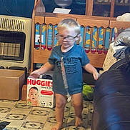 Kohen is registered to the contest to win money with this photo: toddler, child, glasses, blue_shirt, barefoot, indoor, crib, cardboard_box, floor_tile, furniture, living_room, toy, clutter, person, movement, home, couch, box, diaper_box, floor