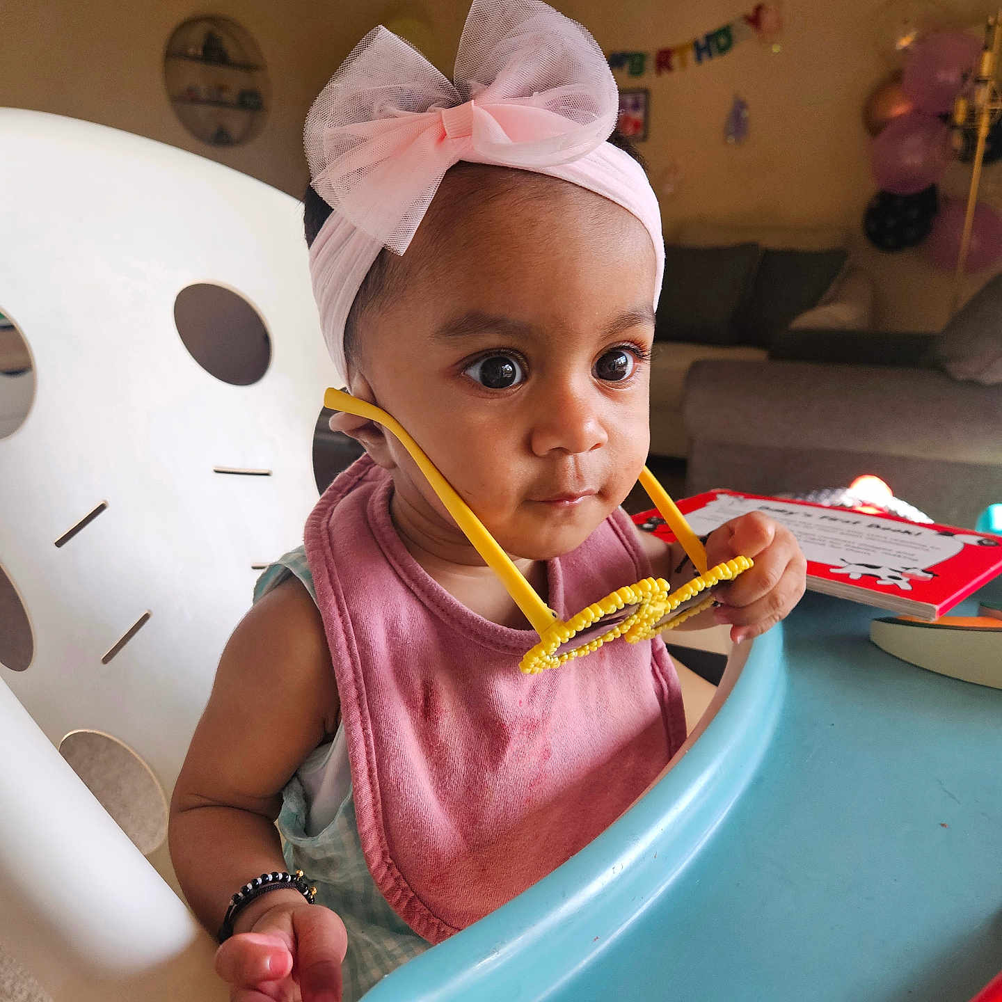 Dhairya joined the competition — help win amazing prizes! baby, child, high_chair, pink_bow, headband, yellow_glasses, curious, indoor, birthday, balloons, banner, living_room, table, toy, infant, clothing, person, face, seated, celebration