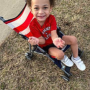 Zayne is registered to the contest to win money with this photo: child, smiling, stroller, red_shirt, curly_hair, outdoor, grass, sneakers, white_shoes, happy, person, casual_clothing, summer, playful, cute, young, portrait, daylight, sidewalk, fun