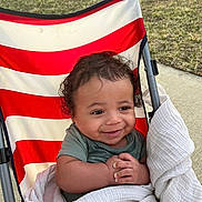 Zavian joined the competition — help win amazing prizes! baby, child, smile, curly_hair, blanket, stroller, outdoor, grass, sidewalk, red_stripes, white_stripes, happy, infant, person, seated, cute, young_child, playful, wrapped, clothing