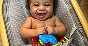 Zavian is registered to the contest to win money with this photo: baby, smiling, curly_hair, toy, baby_seat, patterned_fabric, indoors, happy, child, infant, play, colorful, wooden_frame, flooring, cute, person, skin, sitting, cheerful, young_child