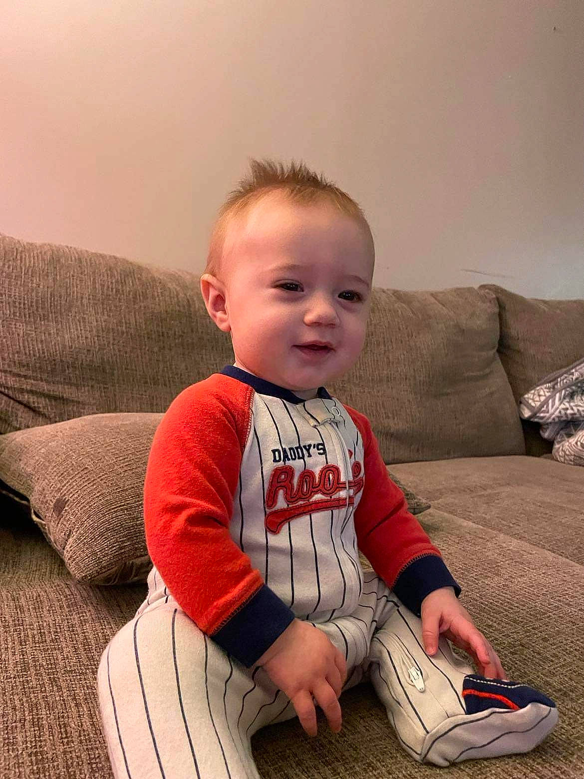 Kayden is registered to the contest to win money with this photo: arm, baby, baby_toddler_clothing, cheek, child, comfort, couch, elbow, flooring, head, human_leg, knee, person, sitting, skin, sleeve, smile, t_shirt, toddler, walking_shoe