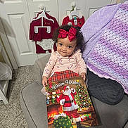 Omyah is registered to the contest to win money with this photo: toddler, child, girl, red_bow, curly_hair, couch, blanket, christmas_book, santa_claus, holiday_outfits, carpet, door, hanger, festive, smiling, indoor, cozy, clothing, portrait, furniture