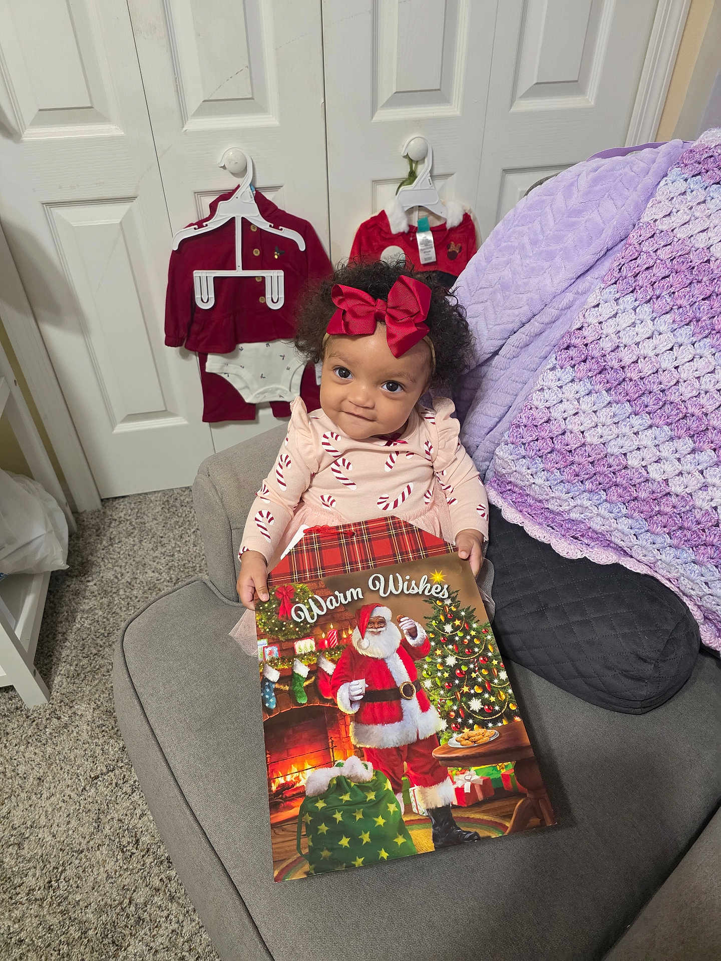 Omyah is registered to the contest to win money with this photo: toddler, child, girl, red_bow, curly_hair, couch, blanket, christmas_book, santa_claus, holiday_outfits, carpet, door, hanger, festive, smiling, indoor, cozy, clothing, portrait, furniture