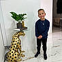 child, boy, smiling, standing, indoor, plant, golden_cheetah, statue, table, books, dartboard, floor, marble_floor, wall, modern, casual_clothing, sneakers, necklace, decor, furniture
