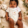 baby, child, smiling, curly_hair, stuffed_animal, bunny, blanket, flowers, floral_arrangement, soft_texture, pastel_colors, onesie, cute, portrait, infant, happy, lying_down, cozy, indoor, skin
