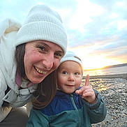 Mateo is registered to the contest to win money with this photo: child, adult, beanie, hat, smile, beach, sunset, sky, sea, water, pebbles, jacket, coat, portrait, closeup, outdoors, family, happy, finger_pointing, person