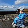 child, toddler, beanie, puffer_jacket, blue_jacket, toy_cart, wheels, sneakers, pavement, boardwalk, bench, waterfront, sea, clouds, sky, outdoors, portrait, sitting, expression, cold_weather