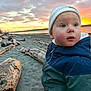Mateo is registered to the contest to win money with this photo: child, toddler, baby, beanie, hat, jacket, beach, sunset, driftwood, sand, ocean, water, sky, clouds, portrait, blue_eyes, rosy_cheeks, warm_light, outdoor, nature