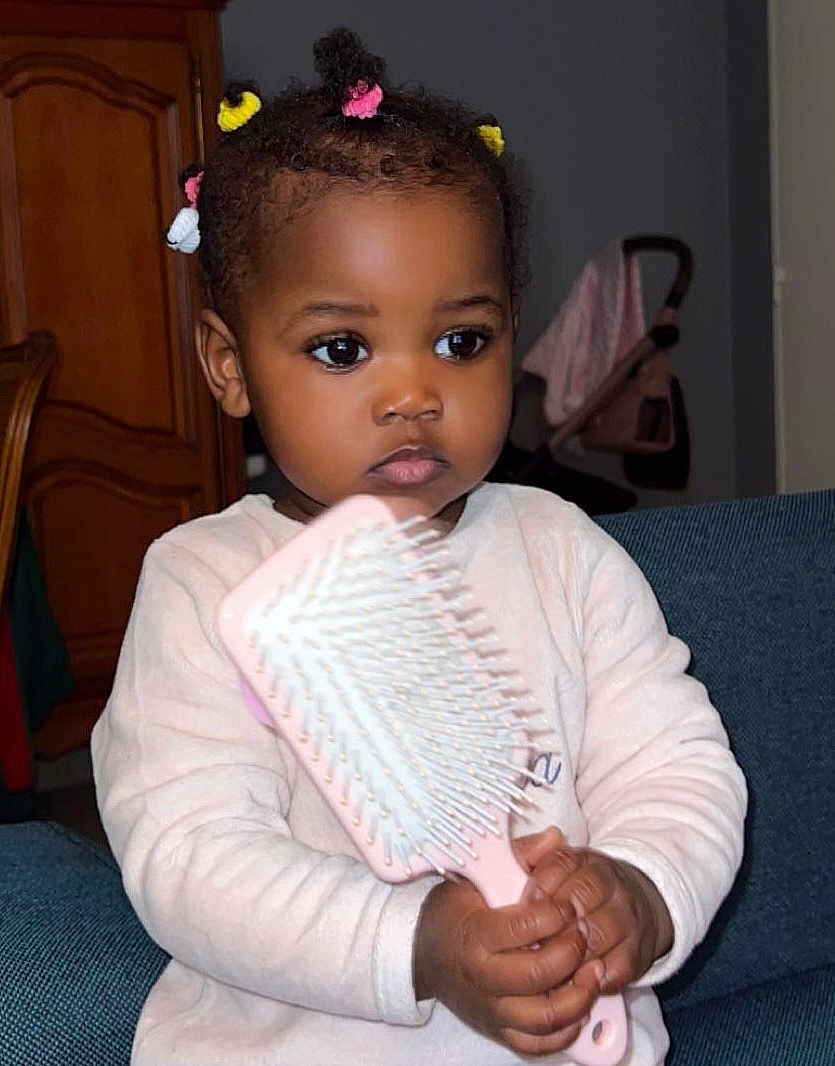 Nehelah participe au concours pour gagner de l'argent avec cette photo : toddler, child, hairbrush, pink, hair_ties, colorful, indoor, sofa, couch, sitting, focused, clothing, long_sleeve, furniture, room, portrait, person, hand, dark_skin, young_child