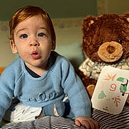 Elio participe au concours pour gagner de l'argent avec cette photo : child, baby, toddler, blue_sweater, stuffed_bear, plush_toy, milestone_card, nine_months, bed, blanket, striped_blanket, portrait, face, big_eyes, surprised_expression, hand, indoor, nursery, cozy, closeup