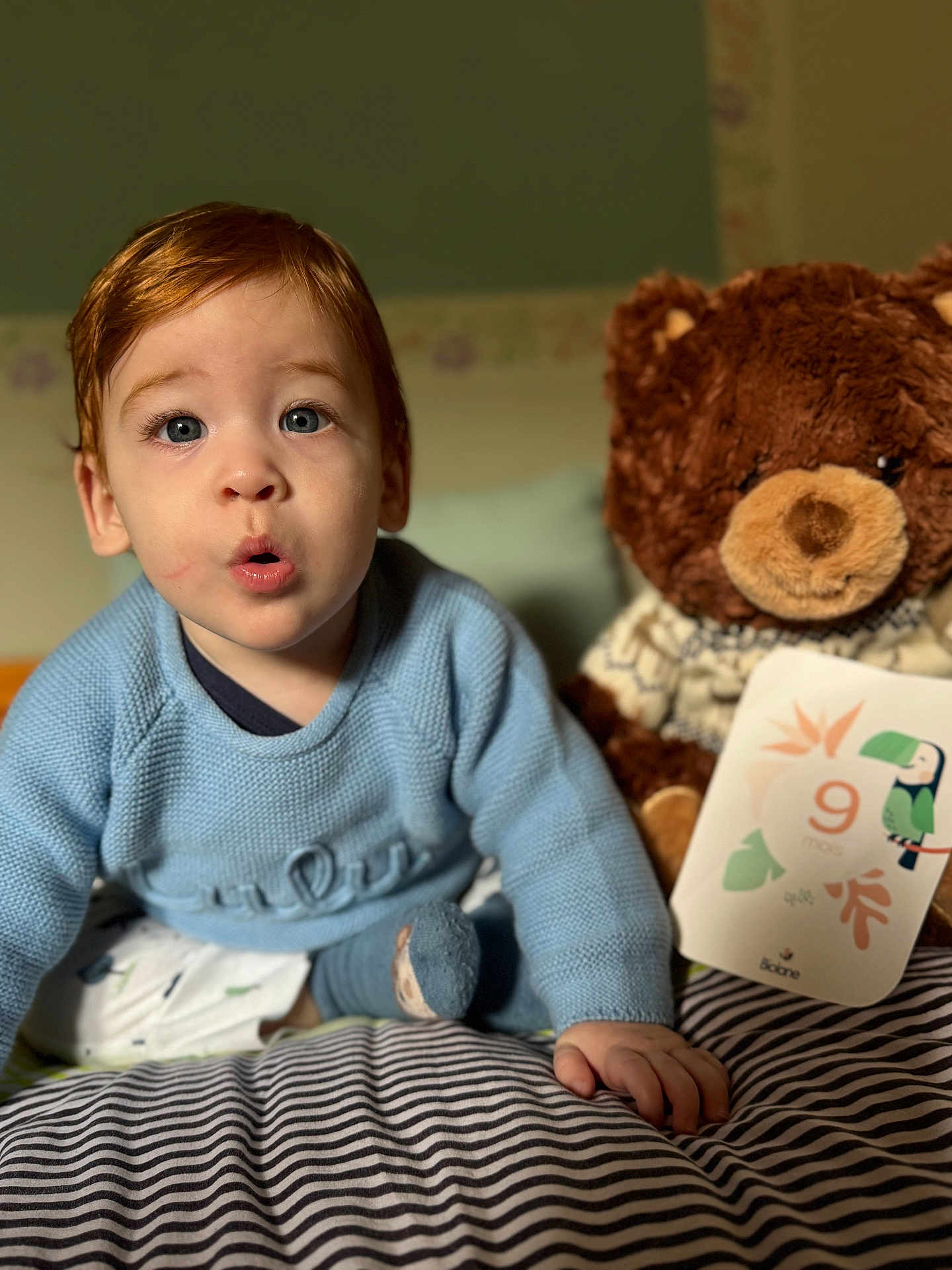 Elio participe au concours pour gagner de l'argent avec cette photo : child, baby, toddler, blue_sweater, stuffed_bear, plush_toy, milestone_card, nine_months, bed, blanket, striped_blanket, portrait, face, big_eyes, surprised_expression, hand, indoor, nursery, cozy, closeup