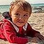 child, toddler, beach, sand, ocean, water, rocks, smile, red_jacket, outdoor, sunlight, happy, portrait, face, hands, casual_clothing, nature, daytime, seaside, young_child