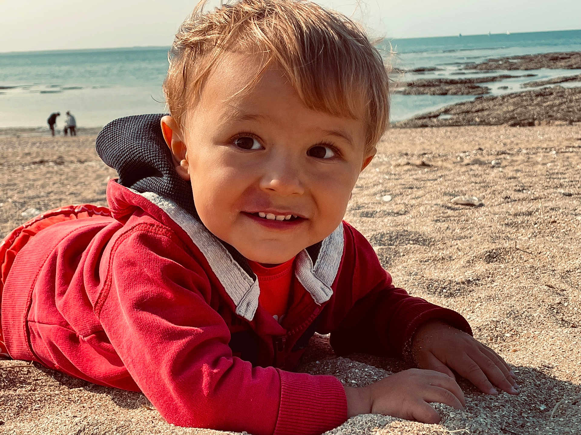 Ruben a rejoint le concours — aidez-le/la à gagner de superbes lots ! child, toddler, beach, sand, ocean, water, rocks, smile, red_jacket, outdoor, sunlight, happy, portrait, face, hands, casual_clothing, nature, daytime, seaside, young_child