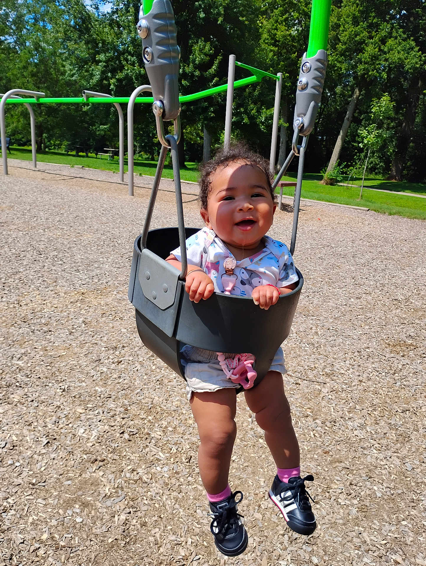Patricia is registered to the contest to win money with this photo: toddler, swing, playground, child, happy, smiling, outdoor, sunny, trees, grass, shoes, shorts, shirt, toy, woodchips, play_equipment, baby, seat, drool, park