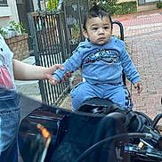 Azrael joined the competition — help win amazing prizes! child, toddler, motorcycle, blue_clothing, outdoor, brick_path, hand_holding, person, fence, plant, window, building, seat, hoodie, pants, short_hair, curious, daylight, sidewalk, casual