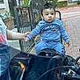 child, toddler, motorcycle, blue_clothing, outdoor, brick_path, hand_holding, person, fence, plant, window, building, seat, hoodie, pants, short_hair, curious, daylight, sidewalk, casual