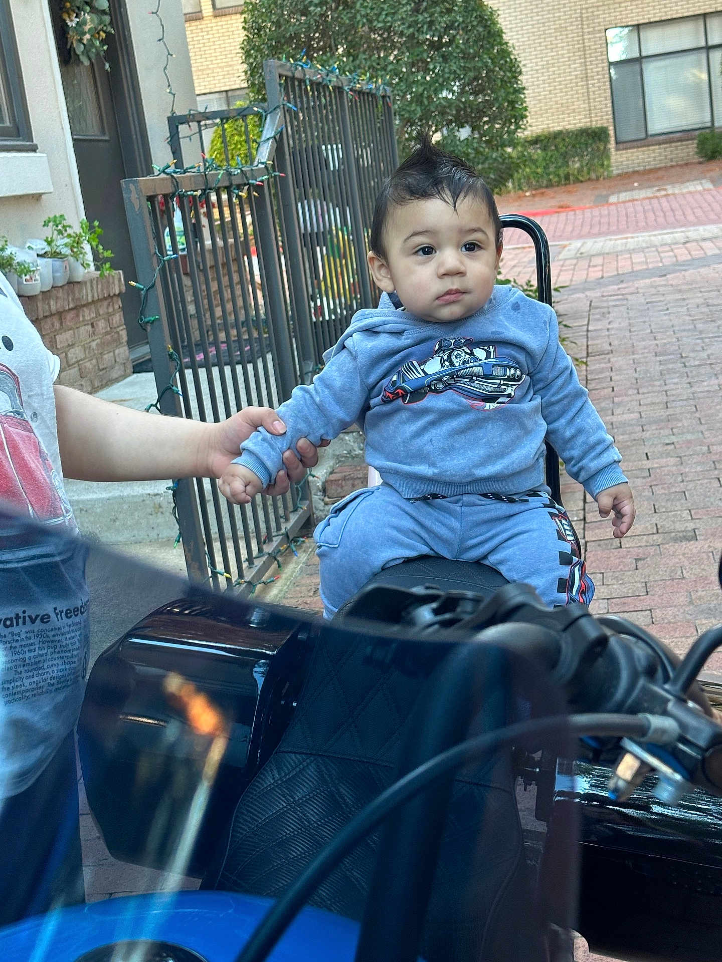 Azrael joined the competition — help win amazing prizes! child, toddler, motorcycle, blue_clothing, outdoor, brick_path, hand_holding, person, fence, plant, window, building, seat, hoodie, pants, short_hair, curious, daylight, sidewalk, casual