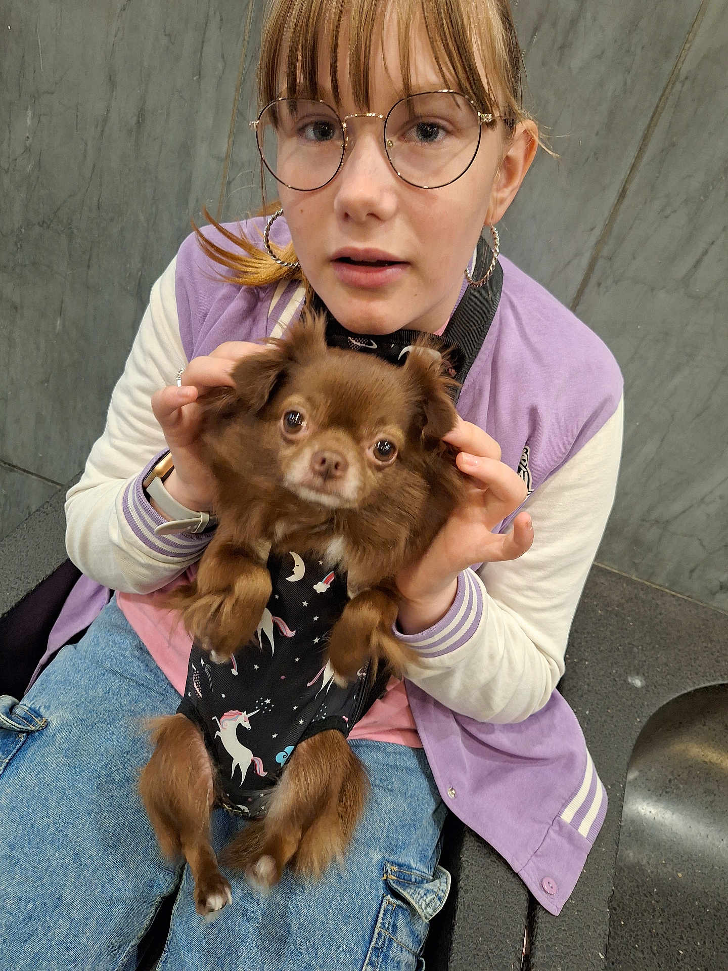 Grisou participe au concours pour gagner de l'argent avec cette photo : person, dog, glasses, jacket, purple, brown_dog, unicorn_print, black_outfit, jeans, sitting, indoor, casual, pet, small_dog, hand, face, expression, wall, bench, earrings