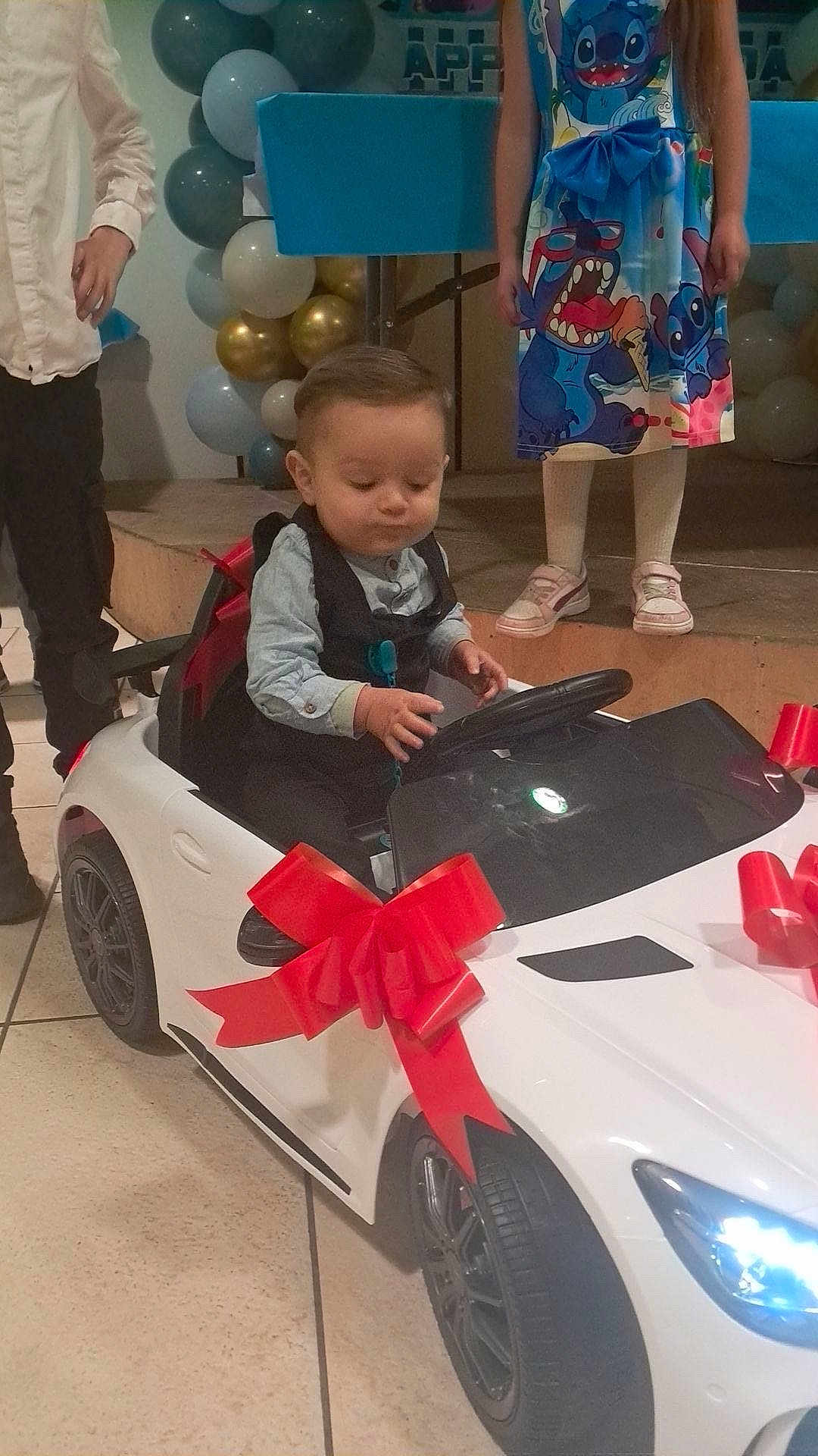 Djuan participe au concours pour gagner de l'argent avec cette photo : toddler, toy_car, red_bow, formal_clothing, child, balloons, party, celebration, indoor, floor_tile, girl, dress, cartoon_print, shoes, steering_wheel, man, stage, event, decorations, people