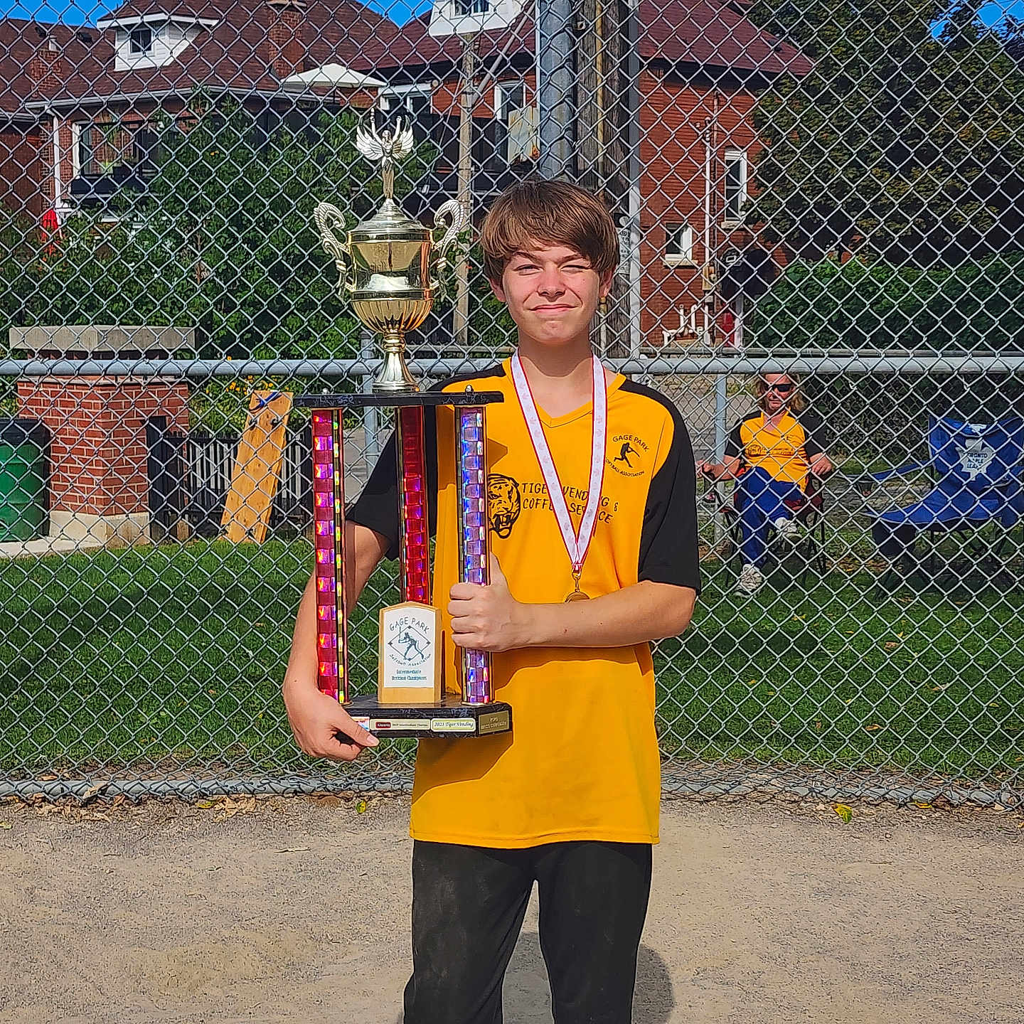 Kayleb joined the competition — help win amazing prizes! child, trophy, medal, sports_uniform, baseball_field, chain_link_fence, sunny_day, blue_sky, grass, dirt, person, victory, pride, spectator, yellow_shirt, black_pants, outdoor, award, celebration, team_sport